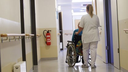 Eine Altenpflegerin schiebt einen Senior im Rollstuhl über einen Flur