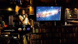 Eine junge Frau (Isabella Rammala) steht mit Mikrofon in einem Pub neben einem Bildschirm, auf dem die Milchstraße zu sehen ist