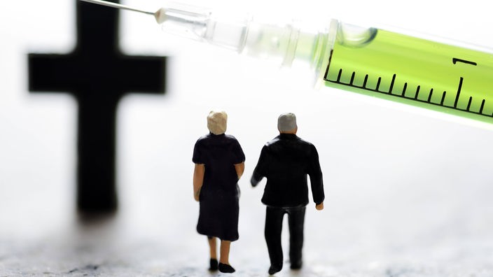 Miniaturfiguren gehen unter einem Kreuz und Spritze - ein Symbolfoto zum Thema Sterbehilfe.