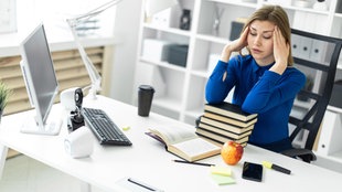 Eine junge Frau sitzt an einem Schreibtisch vor einem Computer und Büchern und hält sich die Hände an die Schläfen