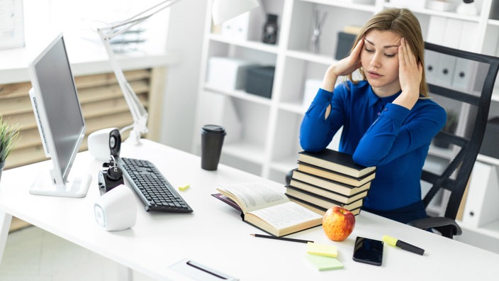 Eine junge Frau sitzt an einem Schreibtisch vor einem Computer und Büchern und hält sich die Hände an die Schläfen