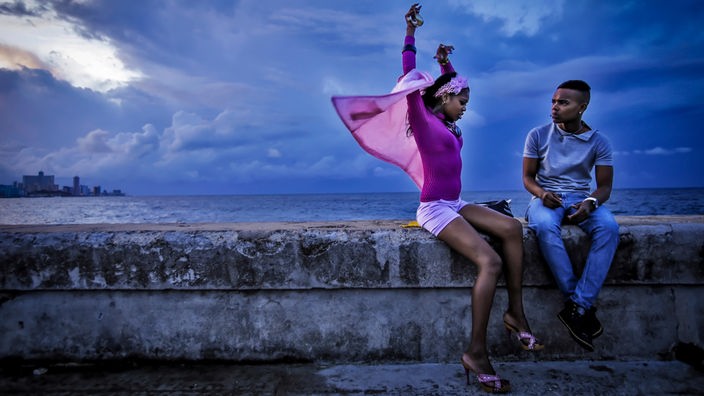 Zwei junge Menschen sitzen in Kuba zur blauen Stunde auf einer Mauer am Meer