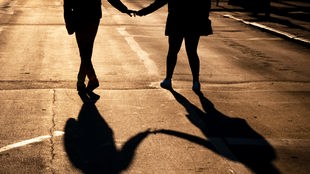 Zwei Menschen, die ihre Hände halten, gehen über einen abendsonnengeflutete Straße, sodass nur ihre Silhoutte und Schatten zu erkennen sind.