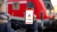 Eine Frau hält ihr Smartphone hoch, sodass das Logo des Deutschlandtickets deutlich auf ihrem Smartphone zu erkennen ist - im Hintergund befindet sich ein roter Zug.