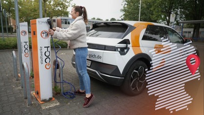 Eine junge Frau mit brauenm Haar, weißer Jacke und Jeans lädt das elektrische Dorfauto der Stadt Goch an einer Ladestation auf.