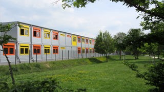 Übergangsbau: graue Container mit gelben und orangefarbenen Fensterrahmen neben einer grünen Wiese, dazwischen ein Zaun