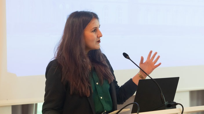 Esther Gardei steht an einem Rednerpult und gestikuliert mit ihrer linken Hand während sie eine Rede hält.