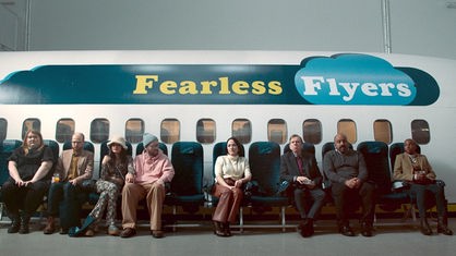Mehrere Menschen Sitzen in einer Reihe auf stühlen vor einem Flugzeug.