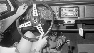 Blick in den Innen- und Fußraum eines Autos im Jahr 1973, Frauenhände am Lenkrad, sie trägt hohe Schuhe und einen kurzen Rock