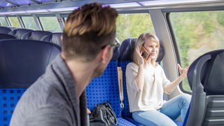 Eine junge Frau telefoniert in einem Regionalexpress mit ihrem Smartphone, während sich ein Mitreisender zu ihr dreht. 