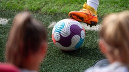 Im Fokus steht ein Fußball der von dem Fußballschuh einer Frau auf dem Rasen gehalten wird, davor sind verschwommen zwei weitere Spielerinnen zu sehen. 