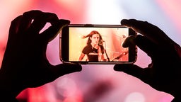 Ein Mensch filmt mit dem Handy eine Frau in einer Metalband auf einer Bühne.
