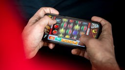 Ein Mensch in eine roten Oberteil hält ein Smartphone in der Hand und spielt virtuelles Glücksspiel.
