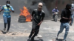 Protestierende Männer rennen in Port-au-Prince über die Straße, im Hintergrund brennen Reifen. 
