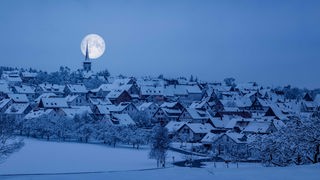 Über einem kleinen winterlich verschneiten Dorf steht im Halbdunkel der Vollmond