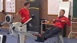 Kai Cziesla auf einer Rudermaschine in einem Trainingsraum, eine Soldatin in Sportkleidung schaut ihm dabei zu