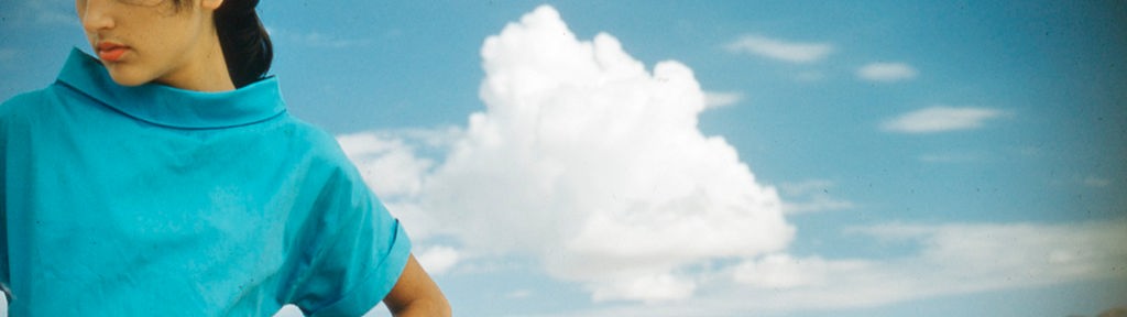 Joan Baez in einem türkis-blauen Oberteil und karierter Hose schauen mit den Händen in die Hüften gestemmt in Richtung Boden, vor blauem Himmel einer Wüstenlandschaft.