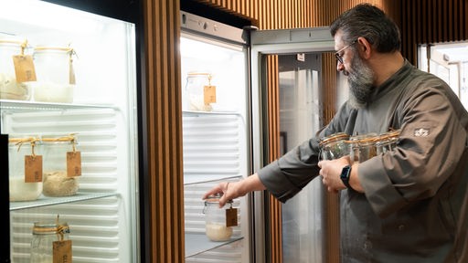 Ein bärtiger Mann räumt Gläser mit Sauerteig in einen großen Kühlschrank.