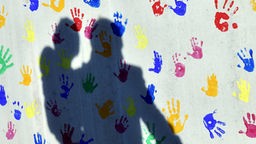 Ein Mann mit einem Kind auf dem Arm und einem an der Hand wirft einen Schatten auf eine mit bunten Handabdrücken bemalte Wand.