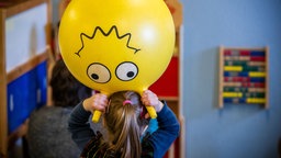 EIn Kind in einer Kindertagesstätte hält einen Hüpf-Ball mit einem gedruckten, lächelndem Gesicht über dem Kopf.