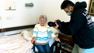 Eine alte Frau sitzt auf einem Bett, daneben steht ein junger Mann in schwarzem Pullover, der eine Kamera hält und filmt.