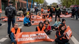Mehrere Mitglieder der Protestbewegung "Letzte Generation" sitzen auf einer Straße und halten orangene Plakate und Spruchbänder in den Händen. 