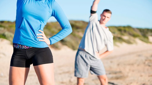 Junge Frau und junger Mann machen Dehnübungen am Strand. 