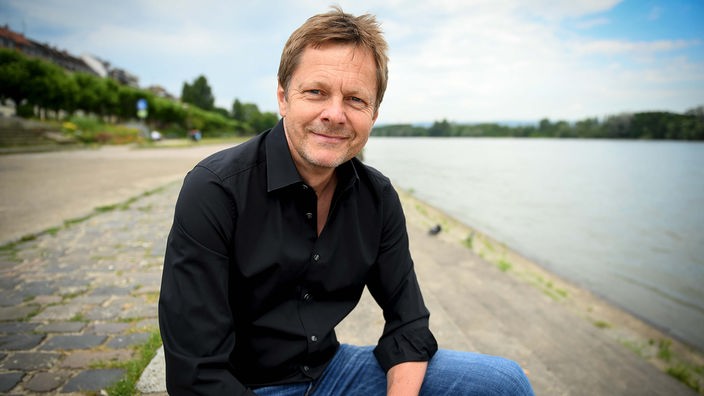 Martin Hecht sitzt am Ufer eines Flusses und lächelt in die Kamera.