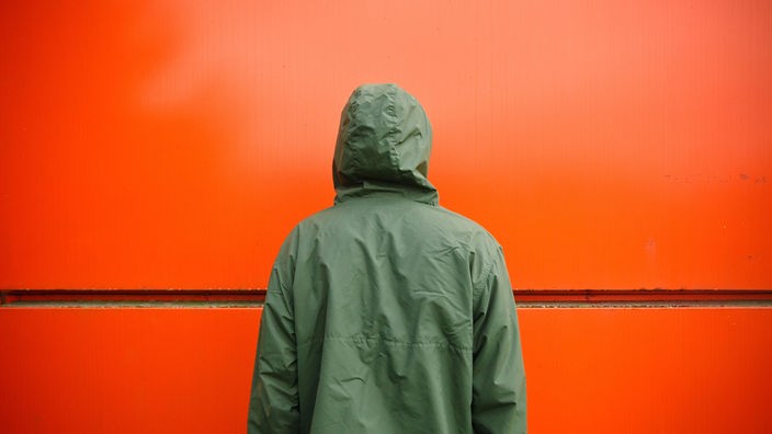 Ein Mann in einer grünen Regenjacke steht mit dem Gesicht zu einer roten Wand