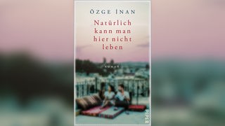Das Buchcover von Özge İnans "Natürlich kann man hier nicht leben" sieht man zwei Menschen auf einem flachen Dach eines weißen Hauses sitzen unter dem Himmel sitzen.