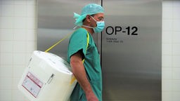 Ein Styropor-Behälter zum Transport von zur Transplantation vorgesehenen Organen wird von einem Mensch in OP-Kleidung am Eingang eines OP-Saales vorbei getragen.