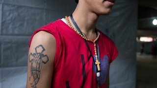 Nahaufnahme auf den Oberkörper eines jungen Mannes der über seinem roten Tanktop einen Rosenkranz trägt und eine ein Tattoo mit christlicher Symbolik auf dem Arm hat.