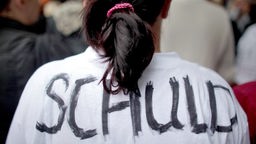 Ein Mensch mit Zopf trägt ein weißes T-Shirt auf dessen Rücken das Wort "Schuld" geschrieben steht.