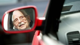 Senior mit Brille sitzt im Auto und blickt in den Außenspiegel.