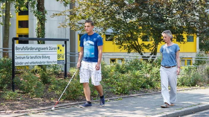 Ein Mann mit Sehbehinderung und einem weißen Stock geht auf einem Bürgersteig