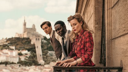 Drei Menschen in Kleidung aus den 1940er Jahren stehen auf einem Balkon und schauen herab.