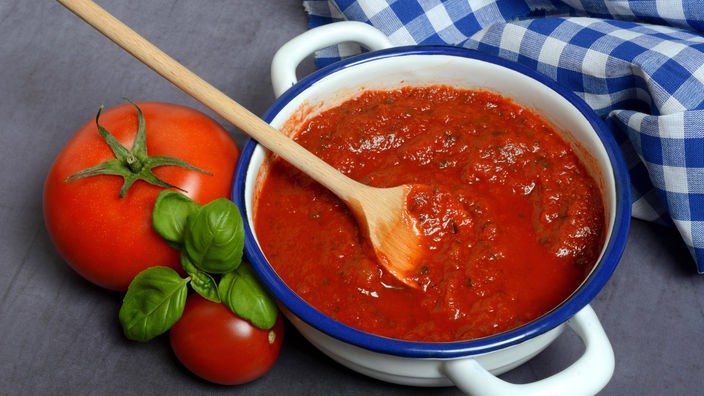 Auf einem Tisch steht neben einigen reifen Tomaten und einem blau-weiß karierten Handtuch eine Schüssel mit Tomaten-Sugo. 