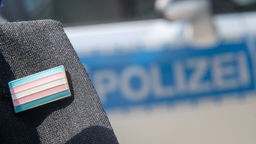 Ein Polizist trägt einen Anstecker der Transgender-Flagge, im Hintergrund ein Polizeiauto