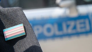 Ein Polizist trägt einen Anstecker der Transgender-Flagge, im Hintergrund ein Polizeiauto