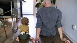 Ein Vater und ein Kind mit Locken und Windel sitzen auf dem Boden - man sieht die beiden von hintern.