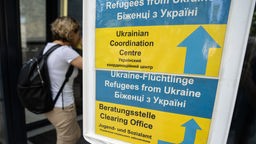 Eine ukrainische Frau an einer Flüchtlingsstelle in Deutschland, daneben ein Schild in ukrainischen Nationalfarben