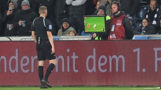 Ein Schiedsrichter steht am Rande eines Fußball-Spielfelds und schaut sich die Wiederholung einer Spielszene auf einem Bildschirm an.