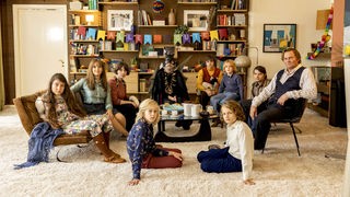 Szene aus "Wann wird es endlich wieder so, wie es nie war?": 10 Menschen in einem Wohnzimmer im Stil der 70er Jahre blicken in die Kamera