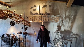 Willi Reiche steht in seiner Maschinenhalle zwischen Kunstmaschinen und unter dem Schriftzug "Art Maschines"