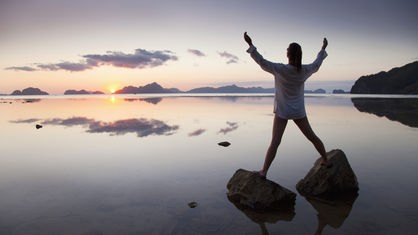 Frau steht mit ausgebreiteten Armen auf zwei Steinen im Meer und blickt in den Sonnenaufgang