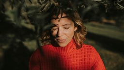 Frau in einem roten Pullover steht unter einem Baum mit geschlossenen Augen