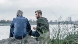 Zwei Menschen sitzen auf Steinen am Wasser und führen ein Gespräch