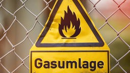 Ein gelbes Warndreieck, auf dem eine Flamme abgebildet ist, hängt an einem Maschendrahtzaun. Darunter steht das Wort "Gasumlage".