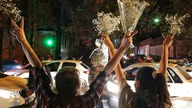 Personen stehen an der Straße und halten Blumenkränze in die Luft. Die Personen stehen mit dem Rücken zur Kamera