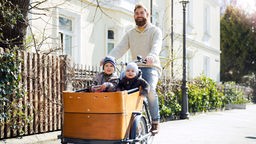 Ein lachender Vater fährt zwei Kinder in eimem Lastenrad durch eine gehobene Wohngegend.